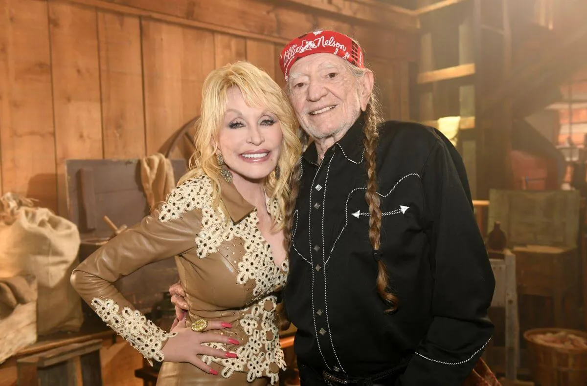 Dolly Parton and Willie Nelson stand with their arms around each other. She wears a brown and white shirt and he wears a black shirt and a red bandanna around his head.