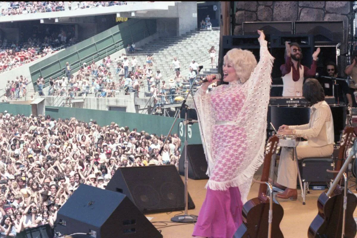 Dolly Parton wears a pink dress with a lace poncho. She stands on stage and sings into a microphone.
