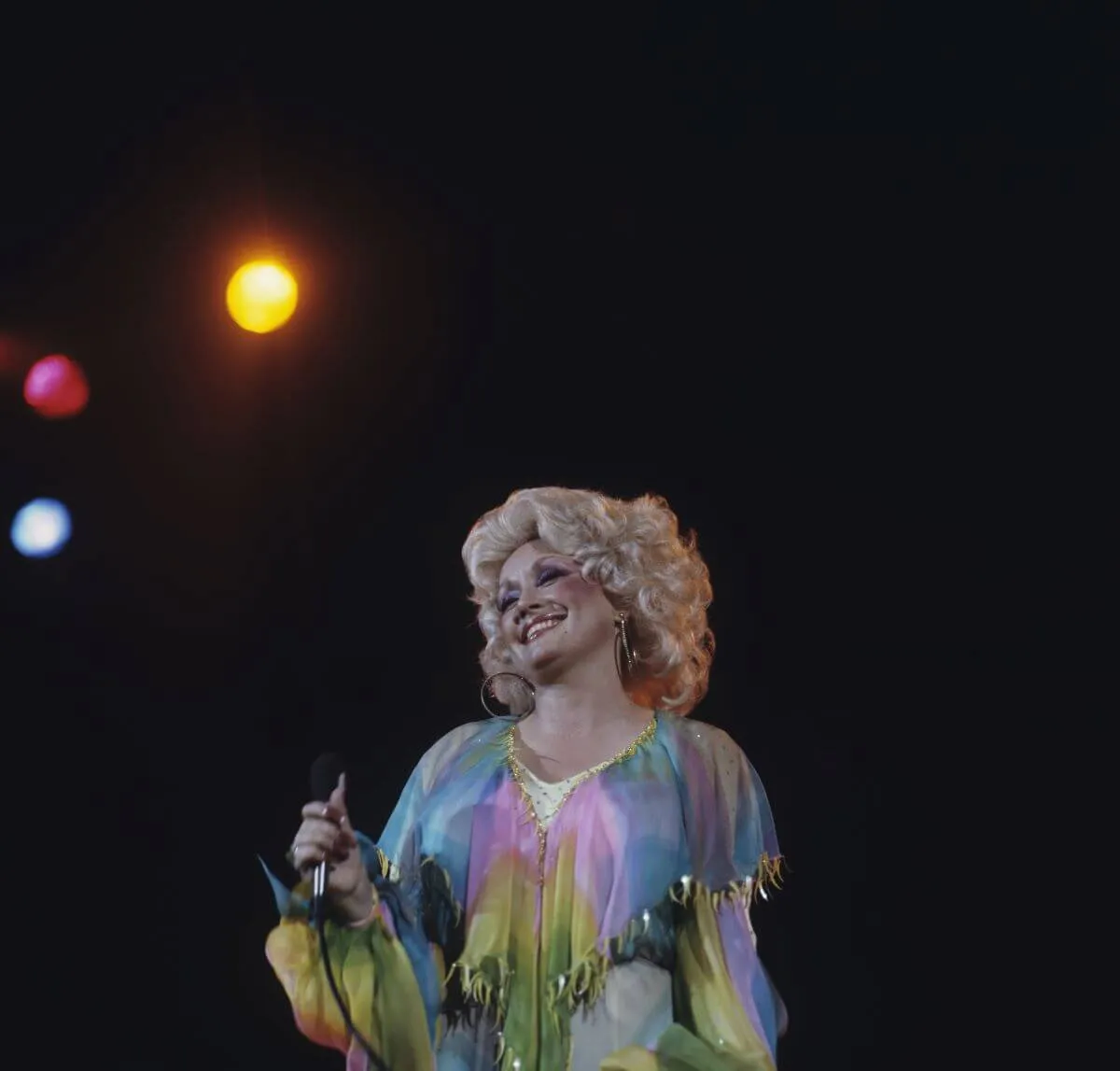 Dolly Parton wears a multicolored shirt and holds a microphone.