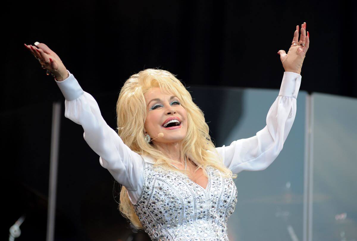 Dolly Parton wears a white shirt with jewels and lifts her arms in the air.
