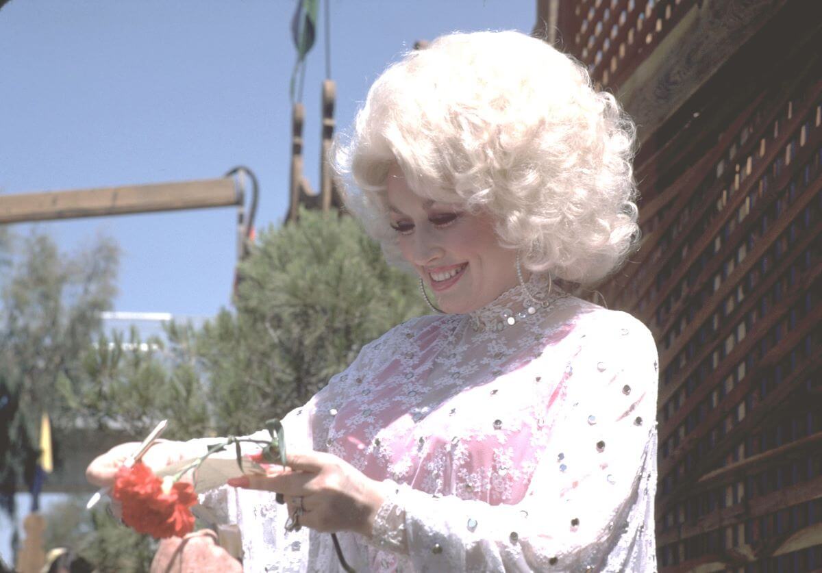 Dolly Parton wears a white lace top and stands outside. She holds a red flower in one hand.