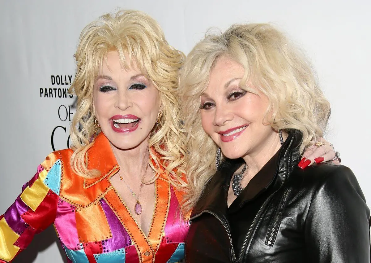 Dolly Parton wears a multicolored coat and stands with her arm around her sister Stella Parton's shoulder. Stella wears a black coat.