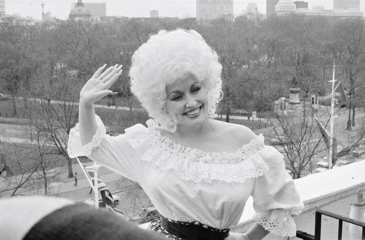 A black and white picture of Dolly Parton standing on a balcony and waving.