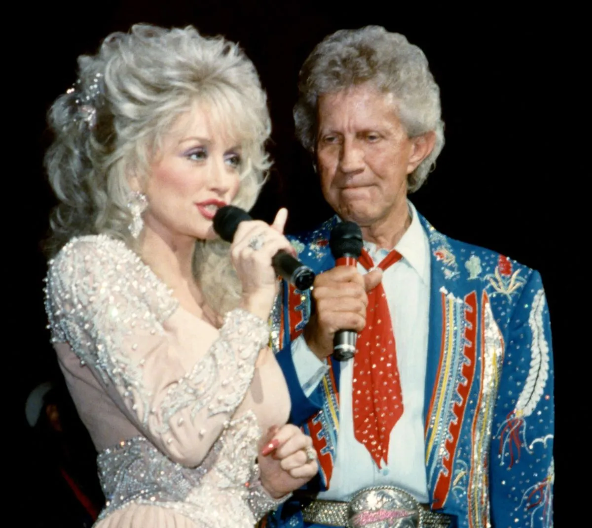 Dolly Parton wears a white dress and speaks into a microphone. Porter Wagoner wears a blue suit and holds a microphone.