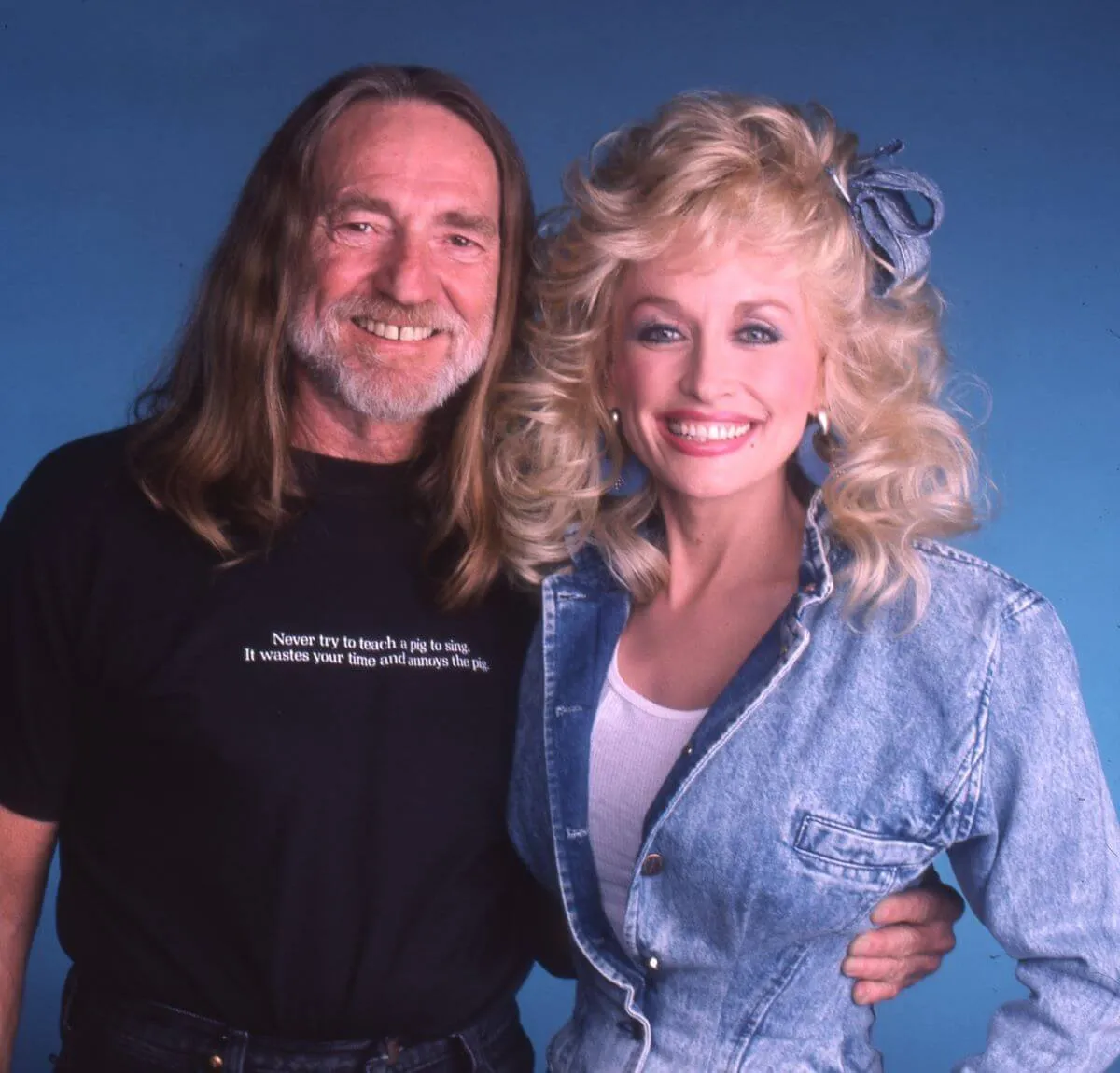 Willie Nelson wears a black shirt and stands with his arm around Dolly Parton's waist. She wears a denim jacket and denim hair bow.