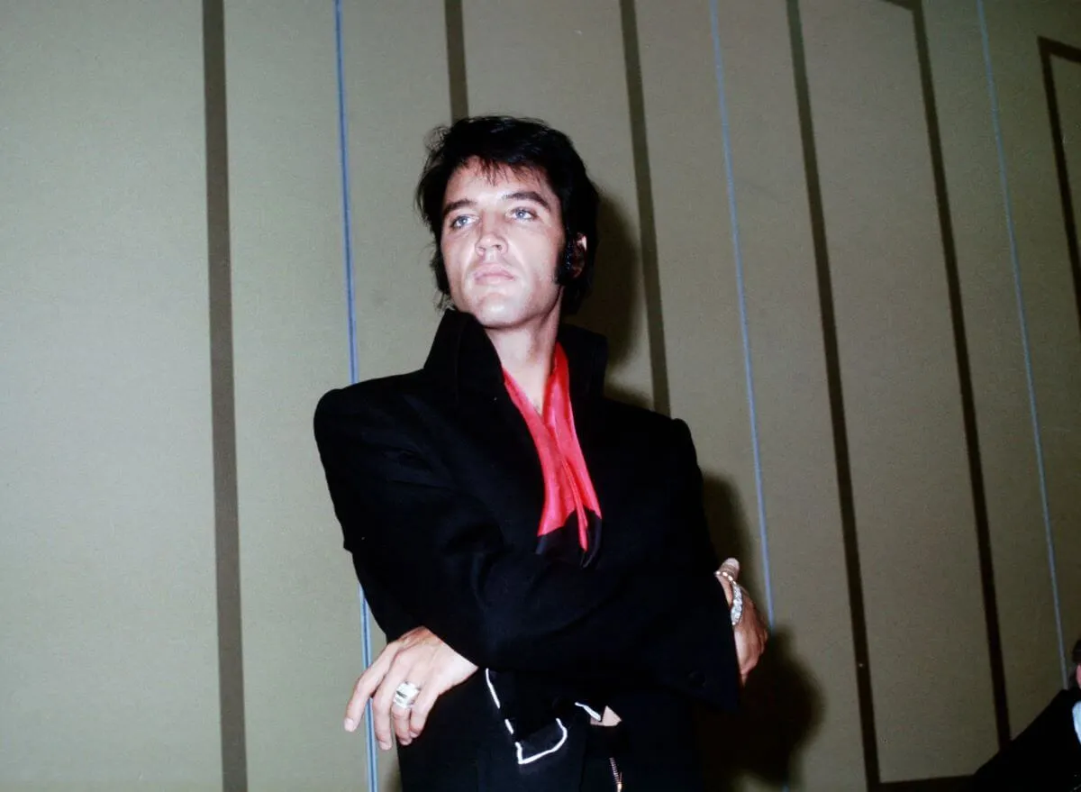 Elvis Presley wears a black suit with a red tie. He stands with his arms crossed.