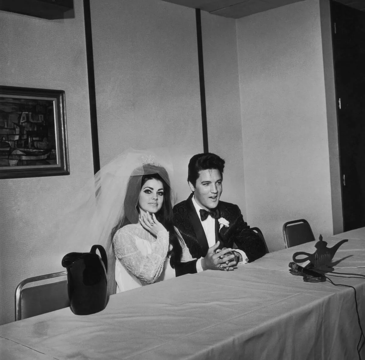 A black and white picture of Priscilla and Elvis Presley sitting on the same side of a long table on their wedding day. She wears a dress and veil and he wears a tuxedo.