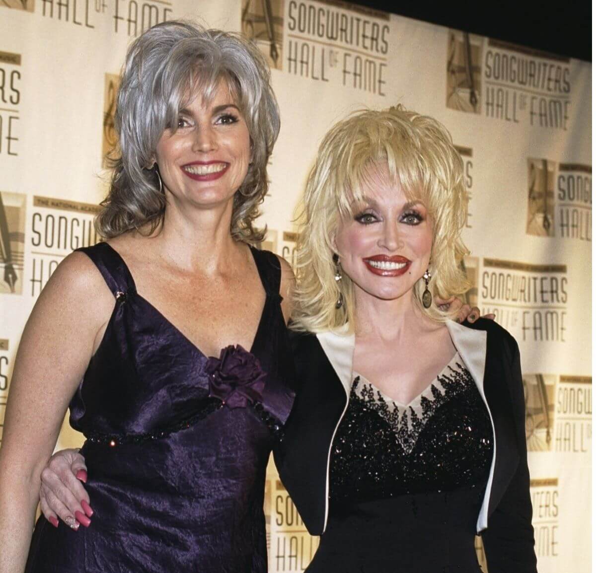 Emmylou Harris and Dolly Parton stand with their arms around each other in front of signage for the Songwriters Hall of Fame. Harris wears a purple dress and Parton wears a black one.