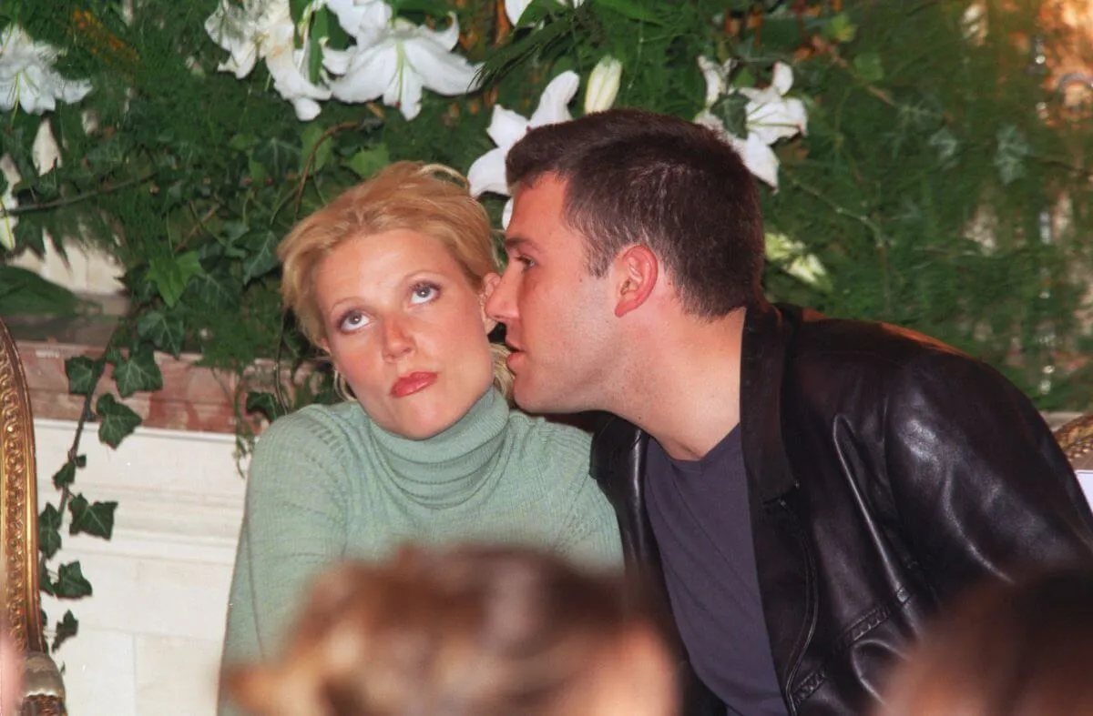 Gwyneth Paltrow wears a green turtleneck and sits next to Ben Affleck, who leans in to whisper in her ear. He wears a leather jacket. She rolls her eyes.