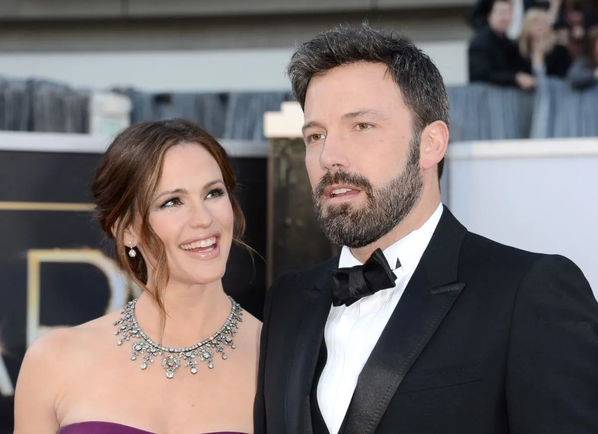 Jennifer Garner and Ben Affleck stand together on a red carpet. She wears a purple strapless dress and a necklace. He wears a tuxedo.