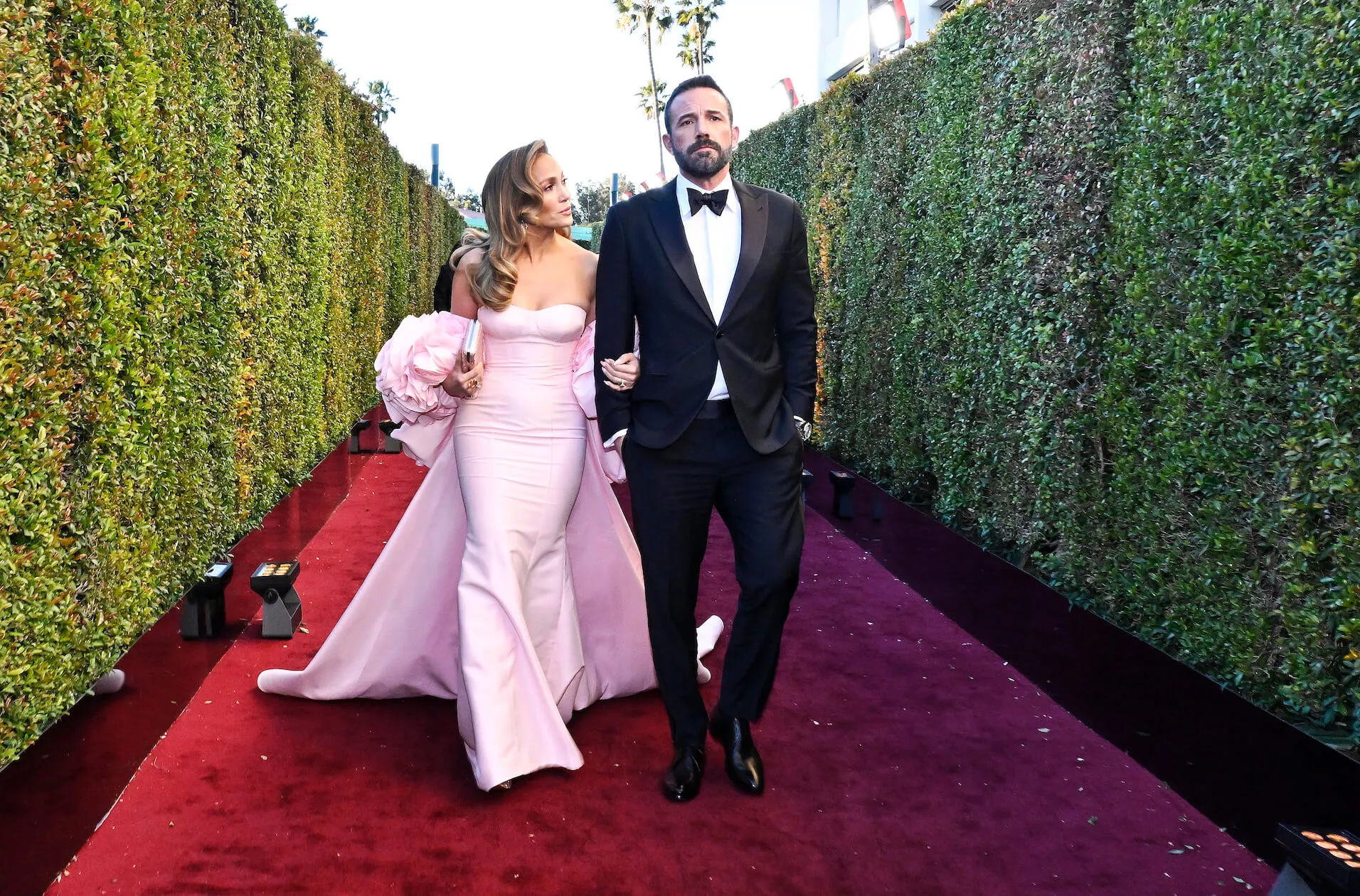 Jennifer Lopez in a pink gown and Ben Affleck in a suit walking on the red carpet between green hedges at the 81st Golden Globes