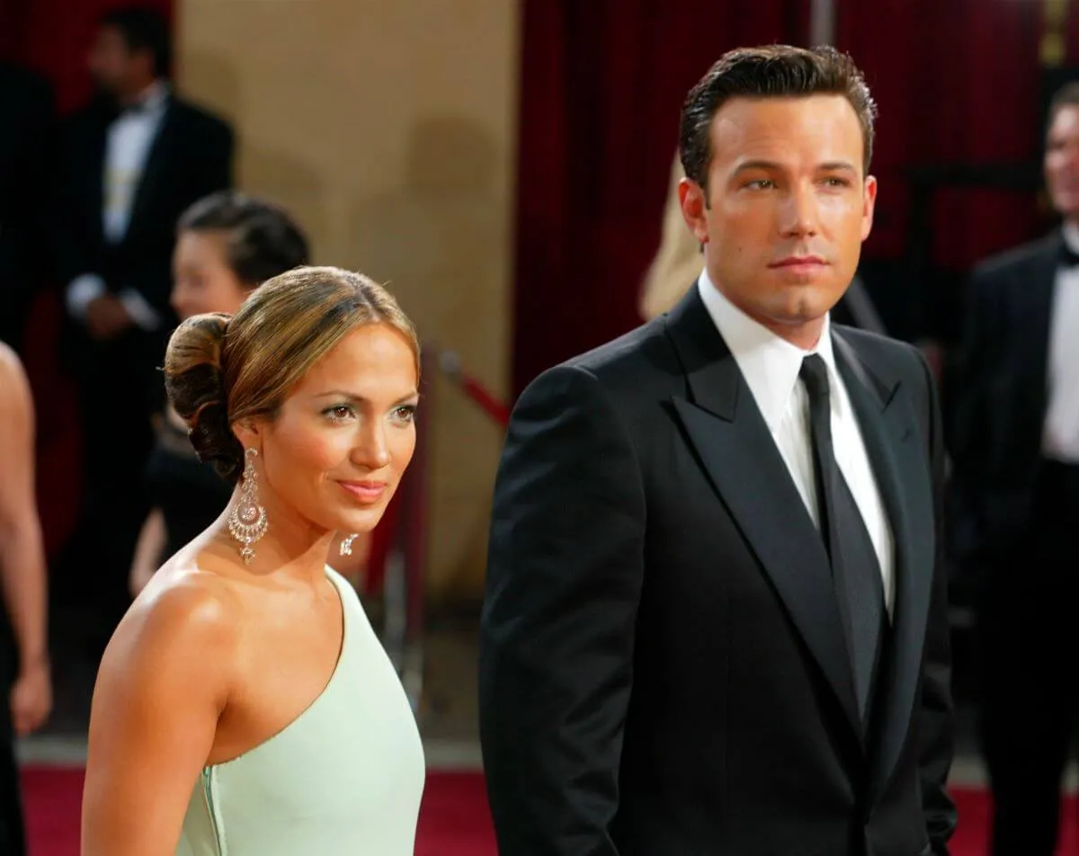 Jennifer Lopez wears a mint colored dress and Ben Affleck wears a suit. They stand on a red carpet.