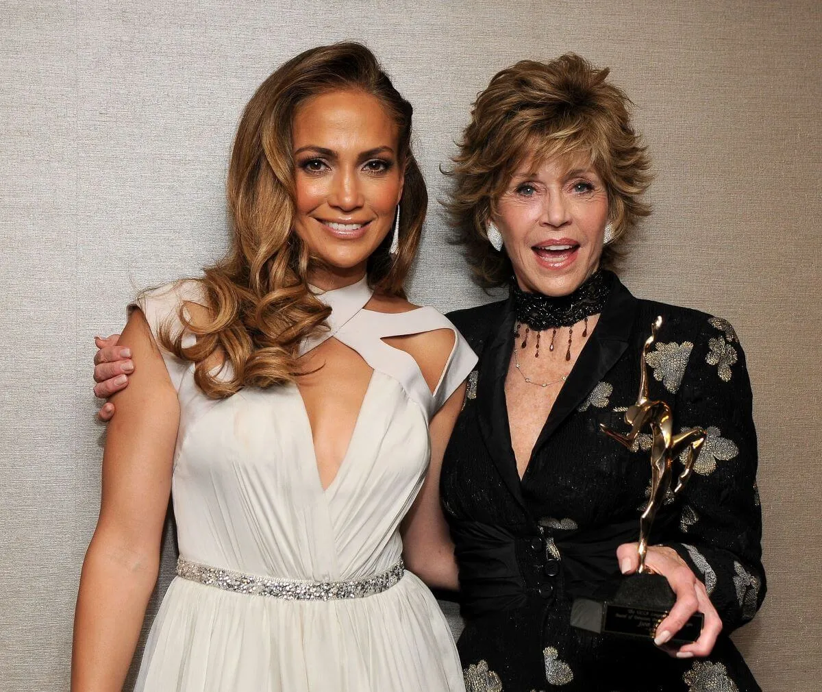 Jennifer Lopez and Jane Fonda stand with their arms around each other. Lopez wears a white dress and Fonda wears a black one.