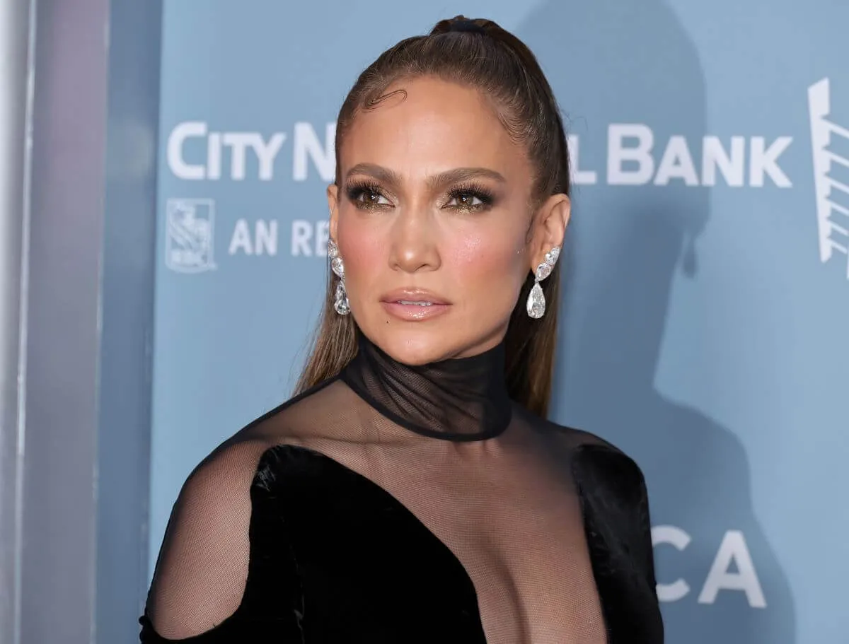 Jennifer Lopez wears a black dress with sheer cutouts. She stands in front of a blue background.