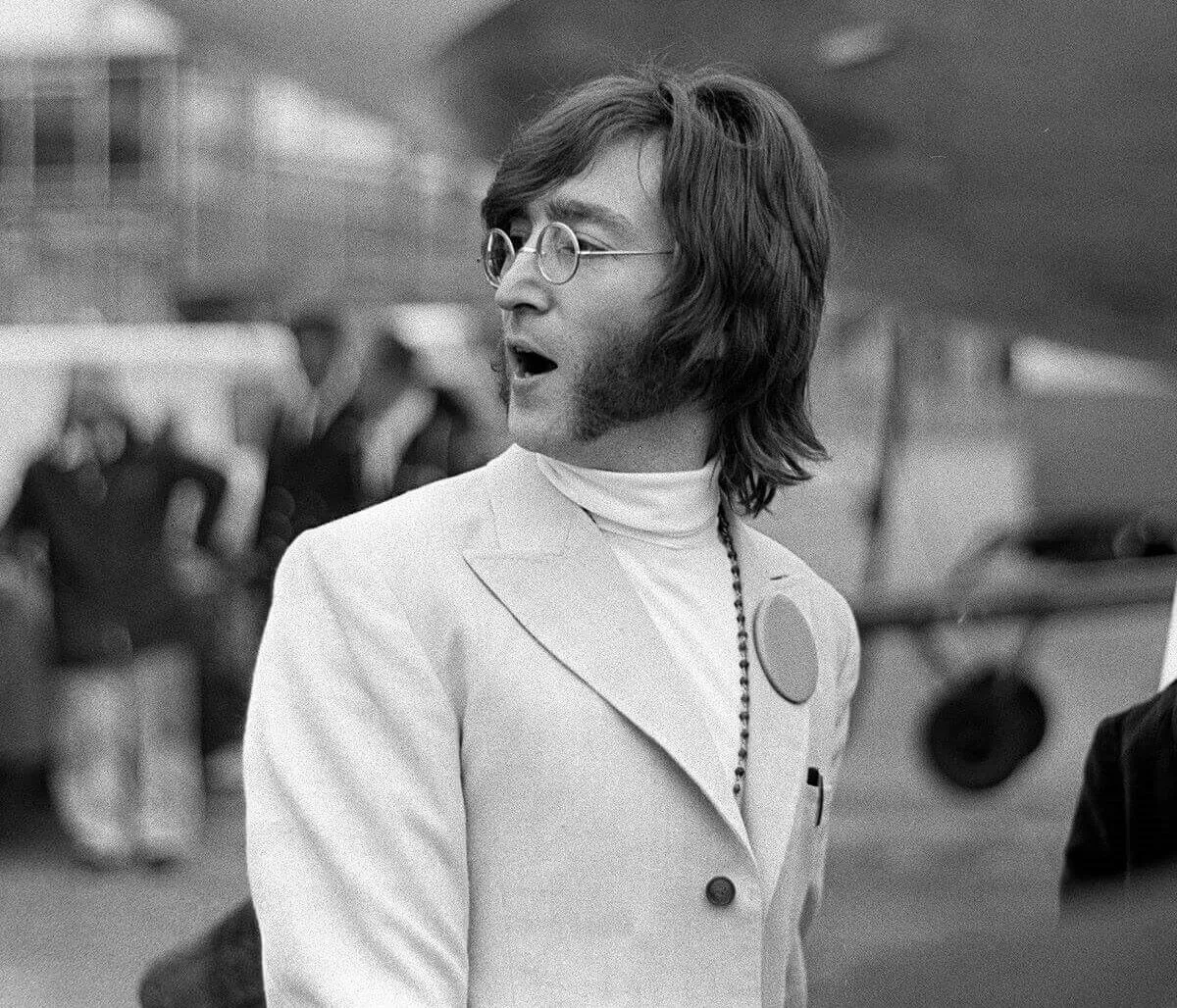 A black and white picture of John Lennon wearing a turtleneck, jacket, and glasses. He stands outside at an airport.