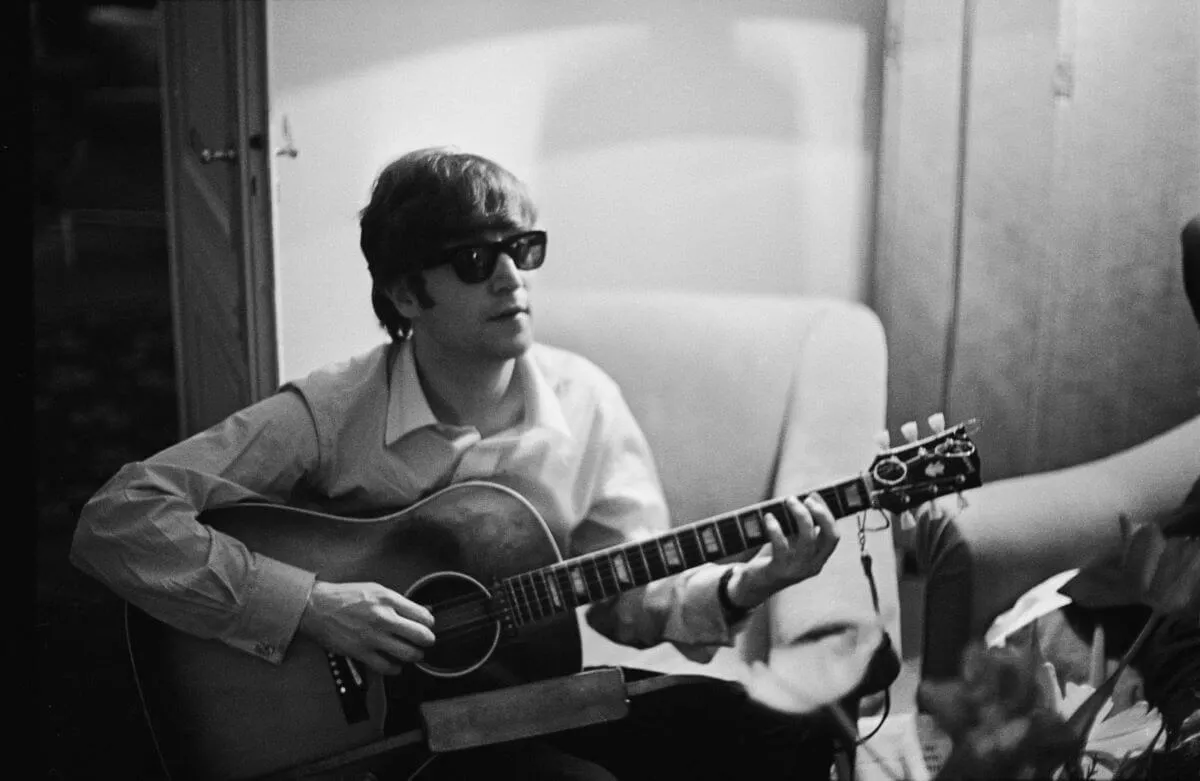 A black and white picture of The Beatles' John Lennon wearing sunglasses and sitting in a chair. He strums an acoustic guitar.