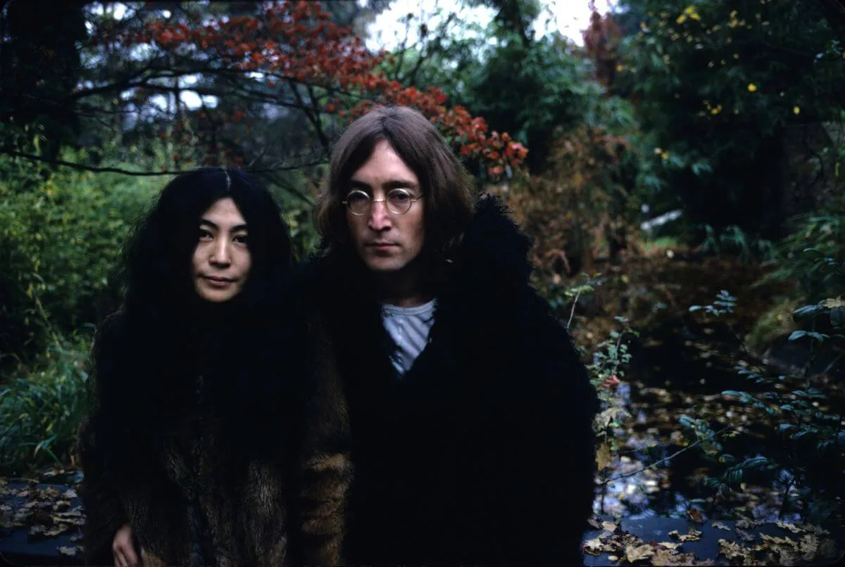 Yoko Ono and John Lennon stand in front of a forested area. They both wear coats.