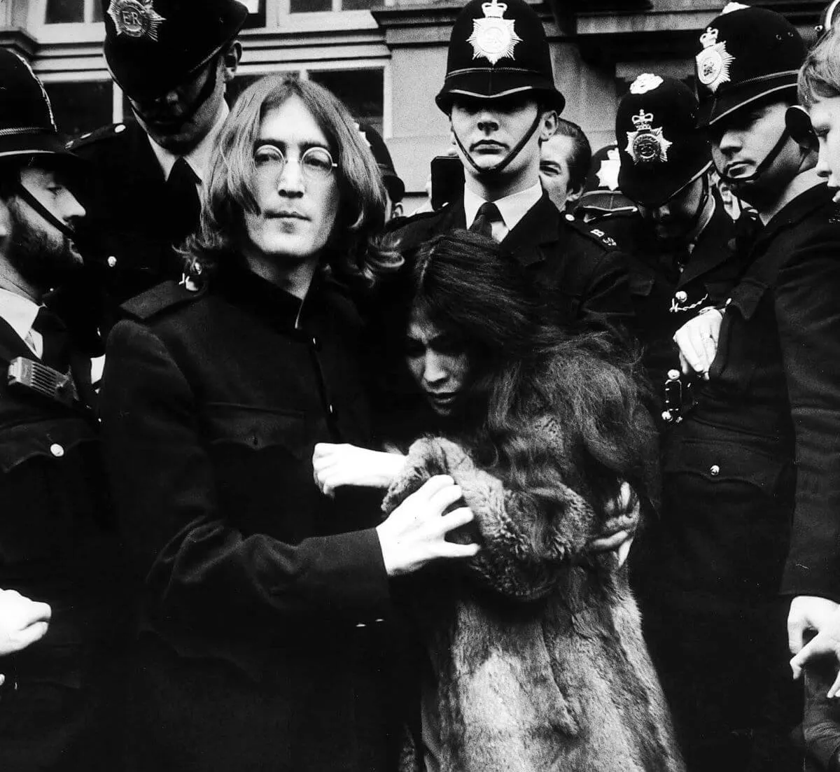 A black and white picture of John Lennon holding onto Yoko Ono. They are surrounded by police officers.