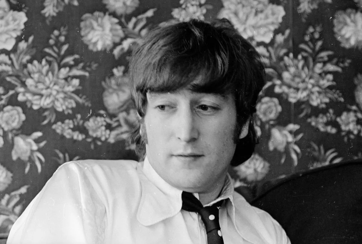 A black and white picture of John Lennon wearing a shirt and tie. He sits in front of a wall with floral wall paper.