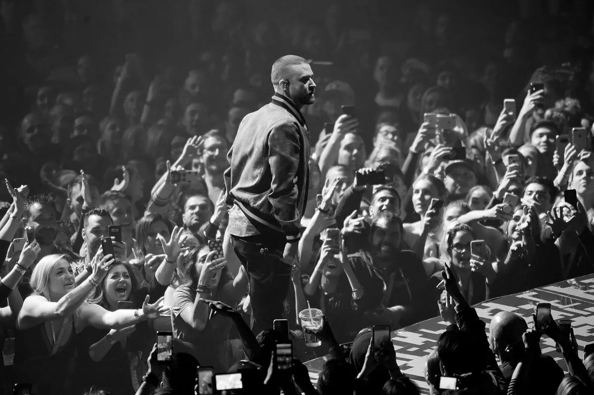A black and white picture of Justin Timberlake standing on a stage in front of fans who reach up toward him.