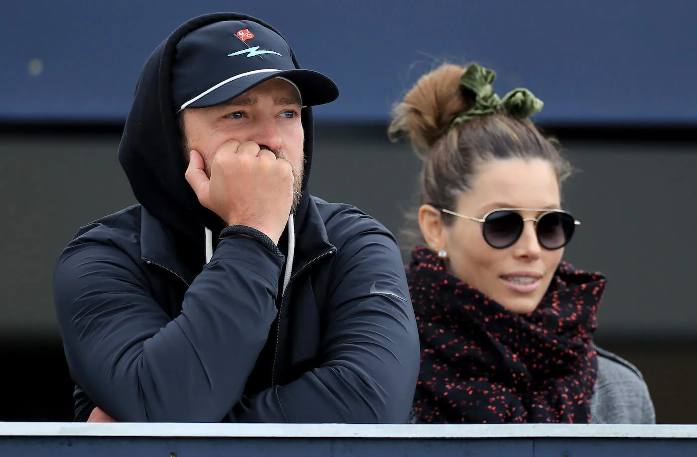 Justin Timberlake resting his head in his hand while wearing a hoodie over his hood. Jessica Biel is next to him in sunglasses and a scarf. The couple is at the Alfred Dunhill Links Championship in 2019.