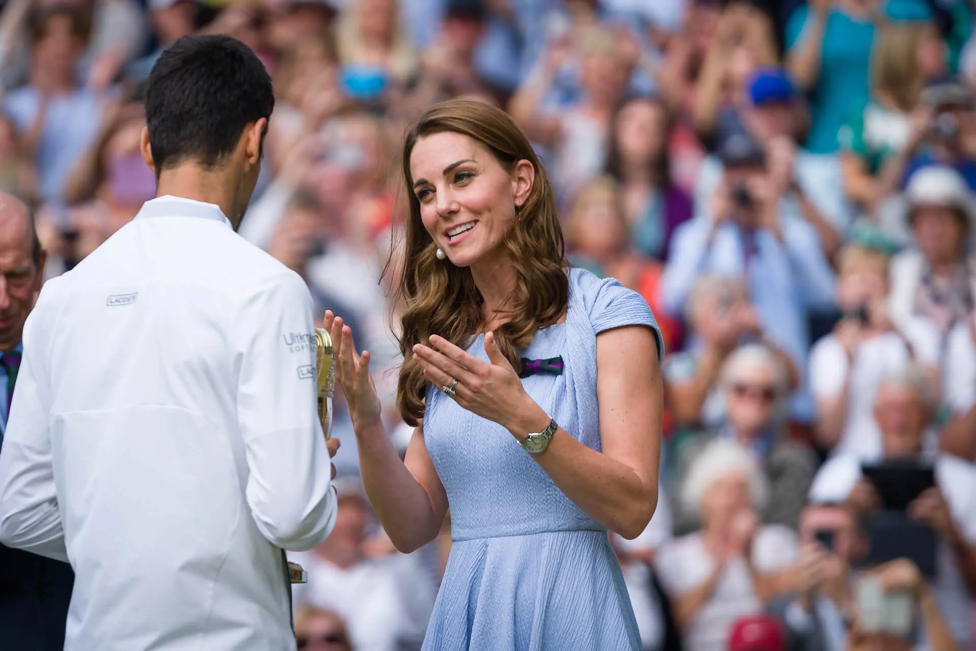 Kate Middleton facing the camera with a crowd behind her and speaking to Novak Djokovic after he wins 