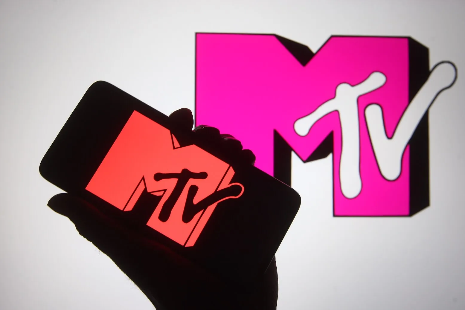 A silhouette hand is seen holding a smartphone with the MTV channel logo on its screen. Another MTV logo is behind it.