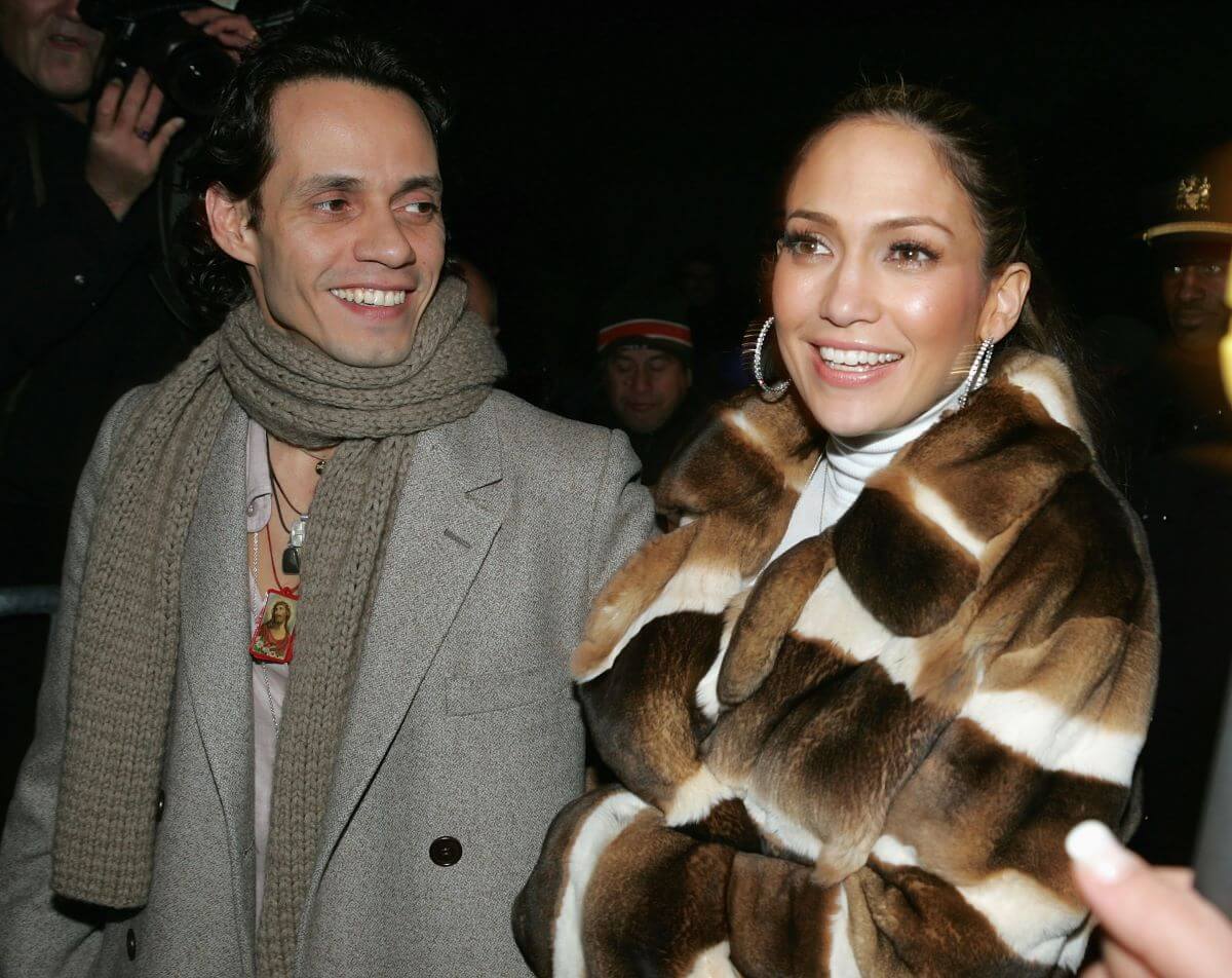 Marc Anthony and Jennifer Lopez wear winter coats and walk through a crowd. They both smile.