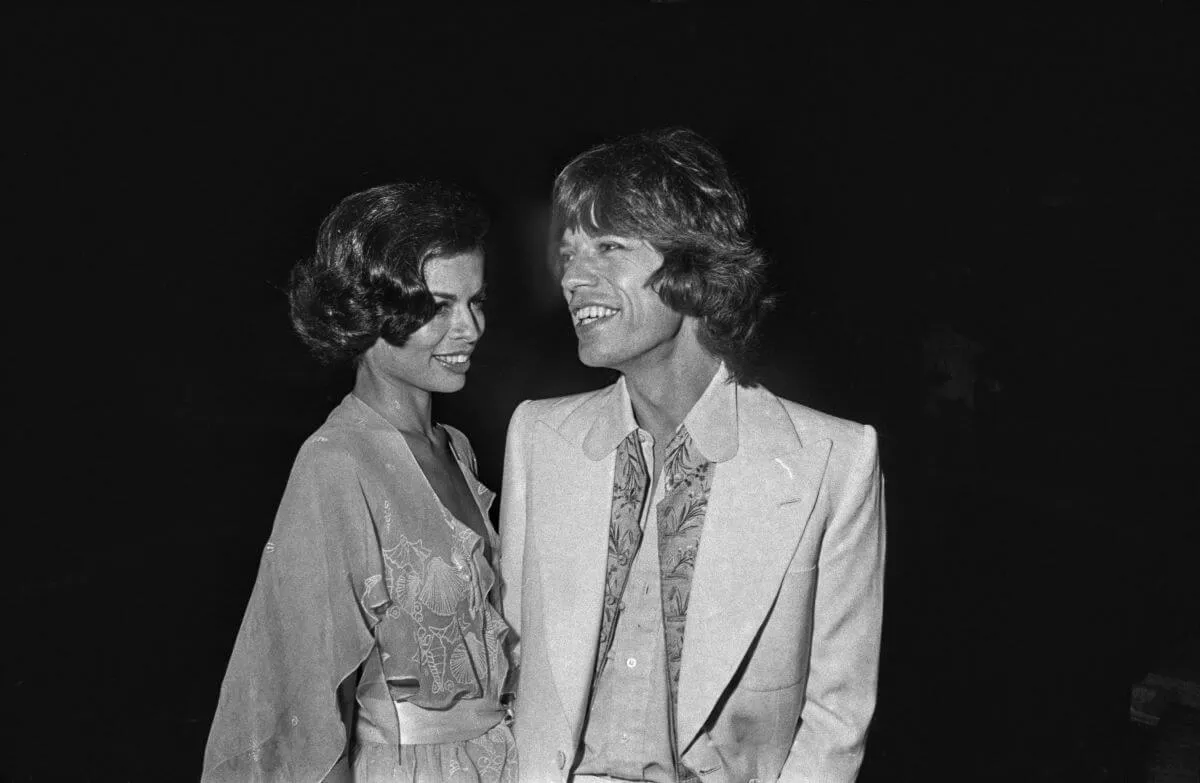 A black and white picture of Bianca and Mick Jagger standing together. She wears a dress and he wears a suit jacket.
