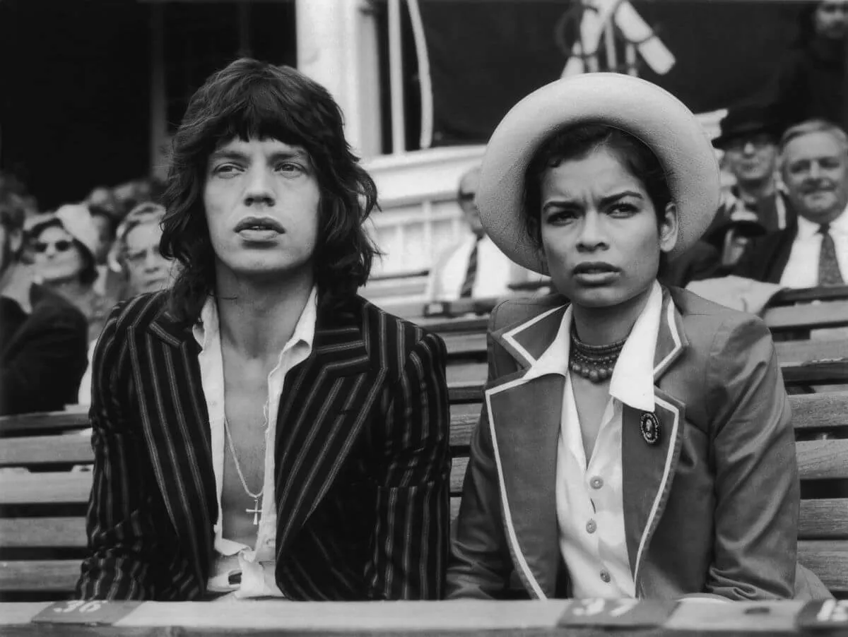 A black and white picture of Mick and Bianca Jagger sitting together. She wears a hat.