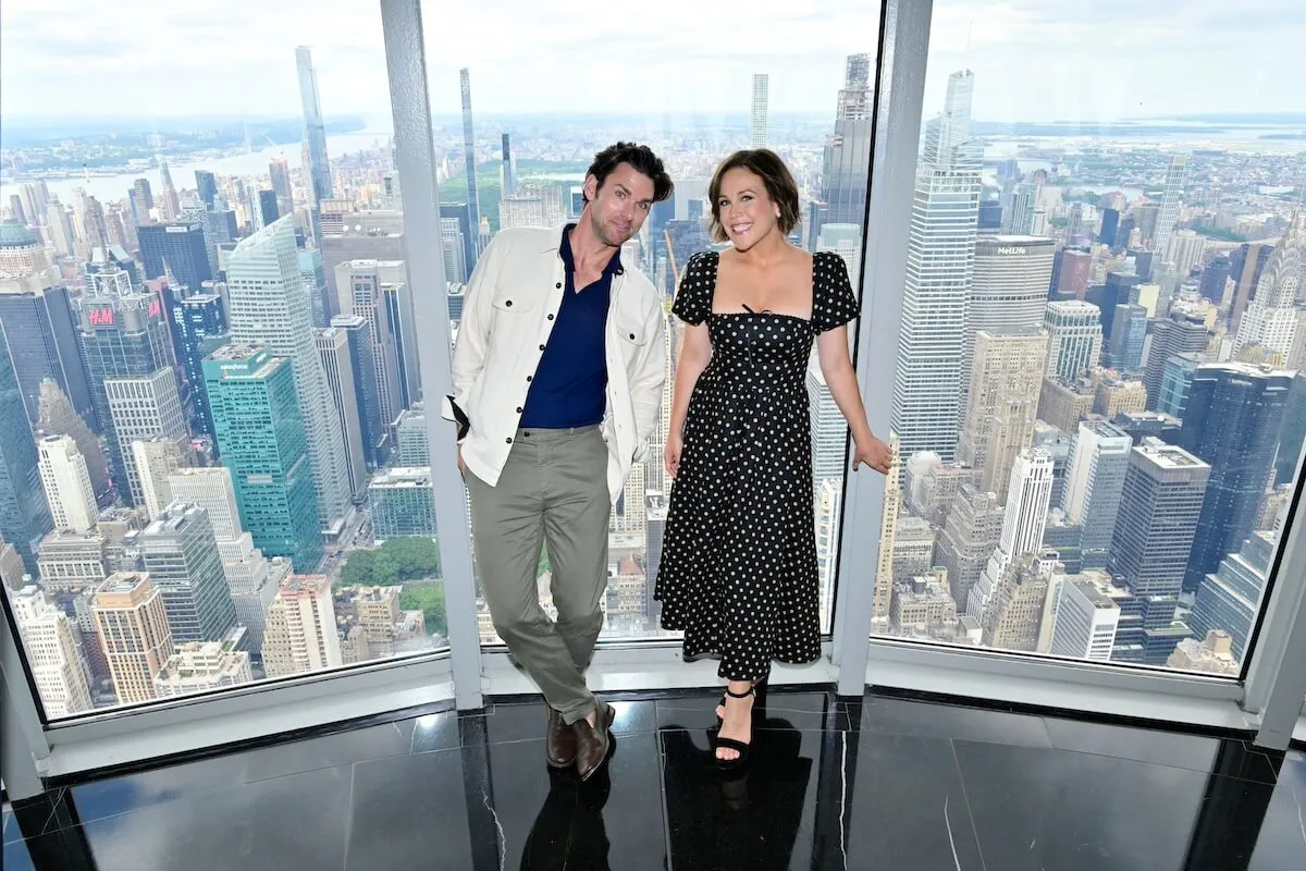 Kevin McGarry and Erin Krakow posing against a wall of windows overlooking NYC