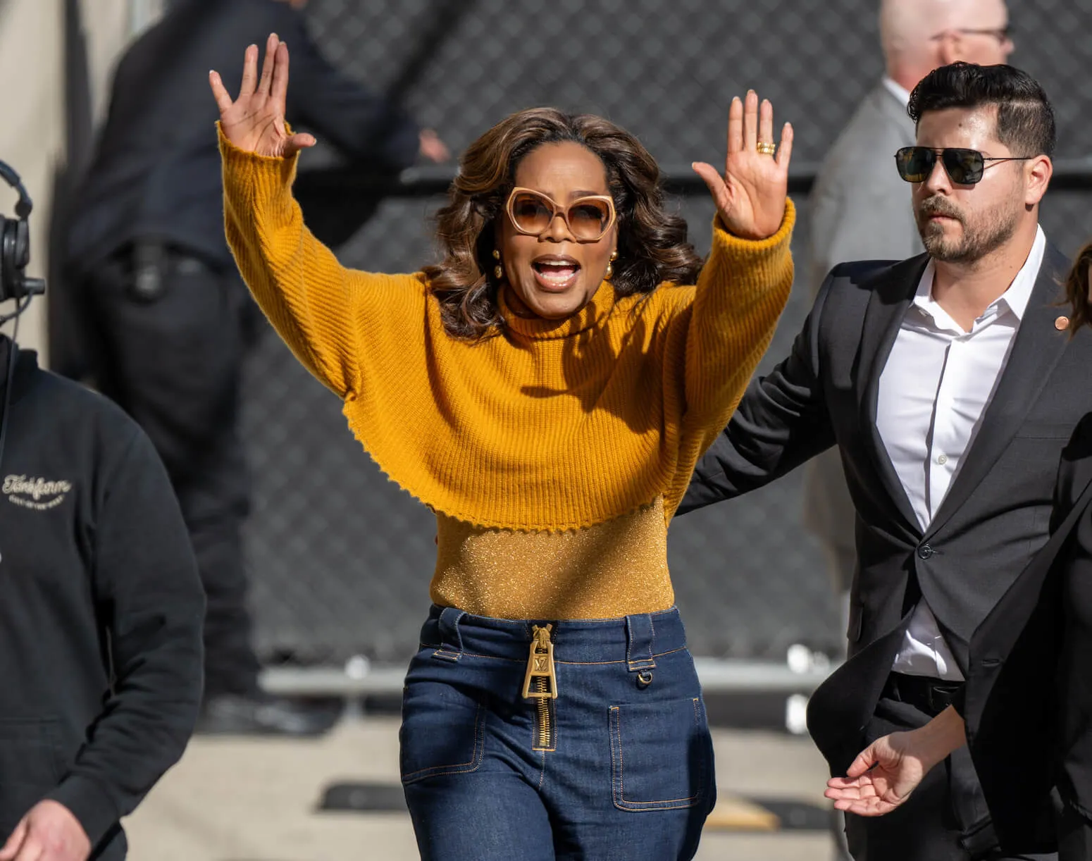Oprah Winfrey holding her hands up while outside. She's wearing a yellow sweater and sunglasses.