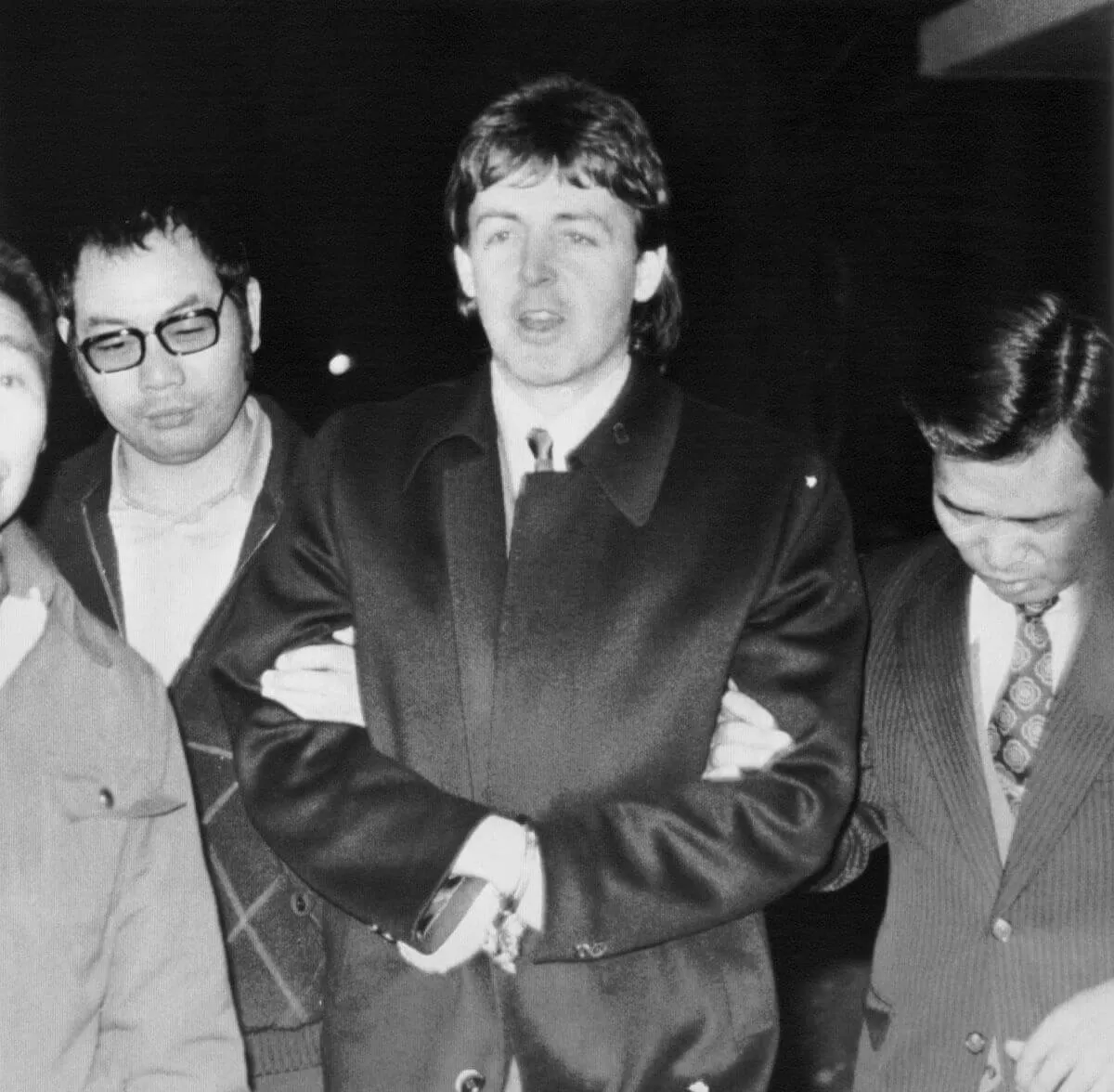 A black and white picture of Paul McCartney being escorted by two men. His hands are handcuffed in front of him.