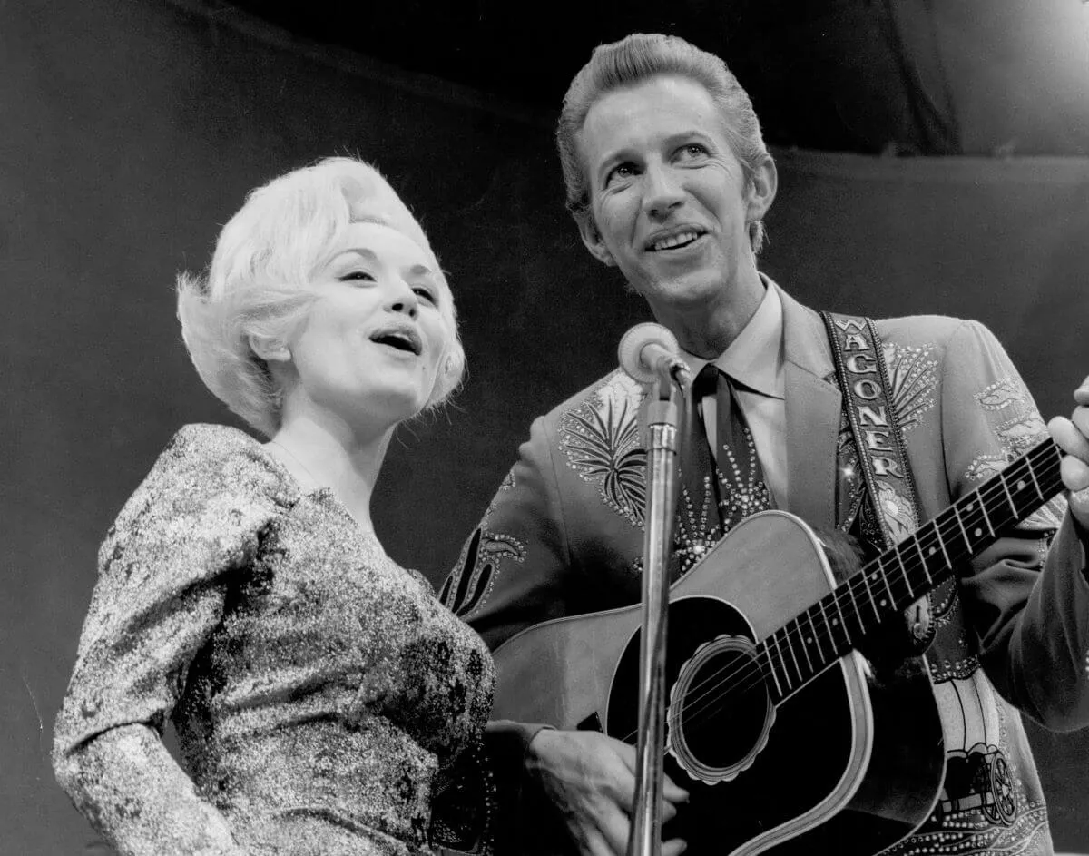 A black and white picture of Dolly Parton and Porter Wagoner singing into the same microphone. He strums a guitar.