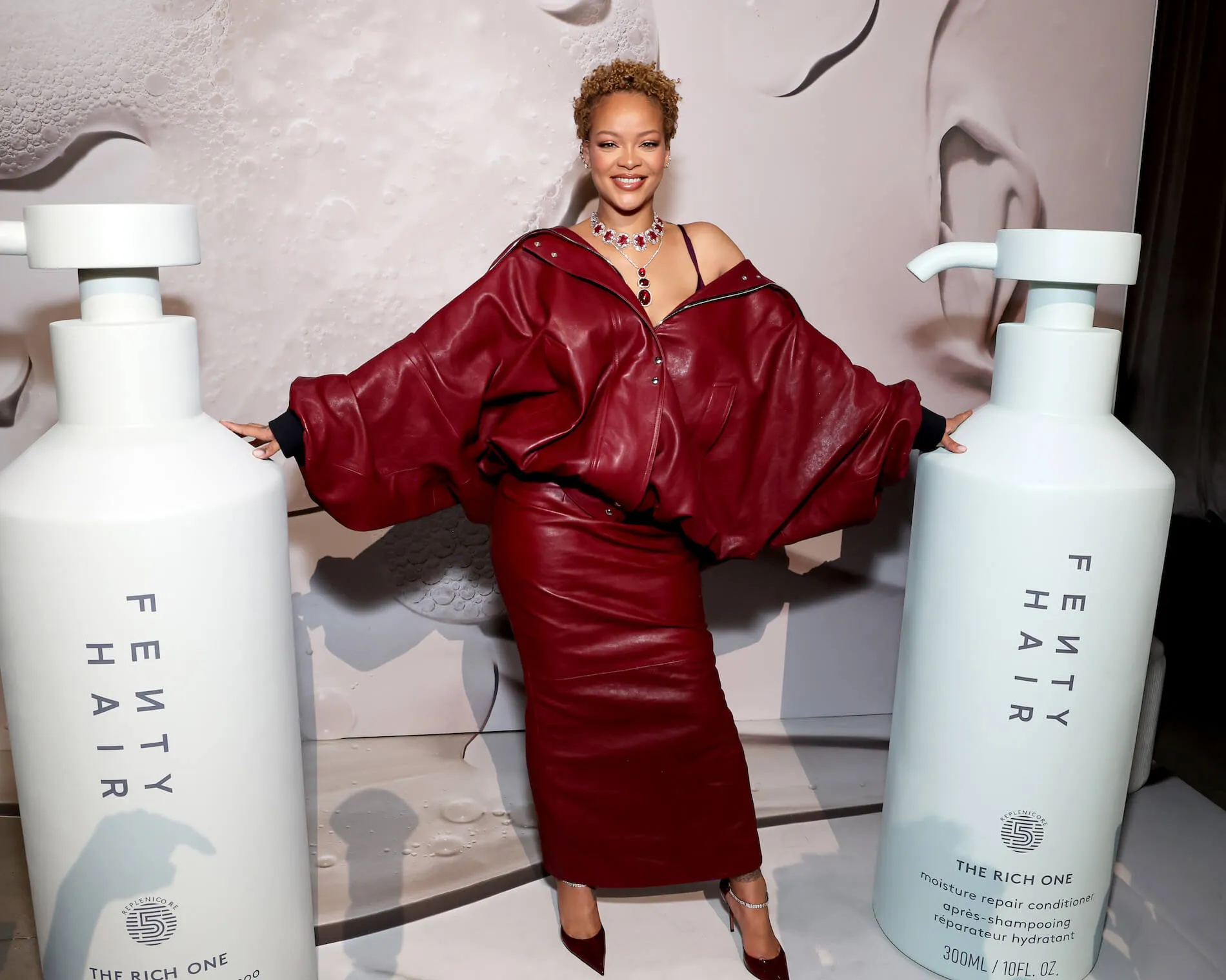 Rihanna posing with natural hair in a red outfit next to two oversized Fenty Hair bottles at the Fenty Hair launch party