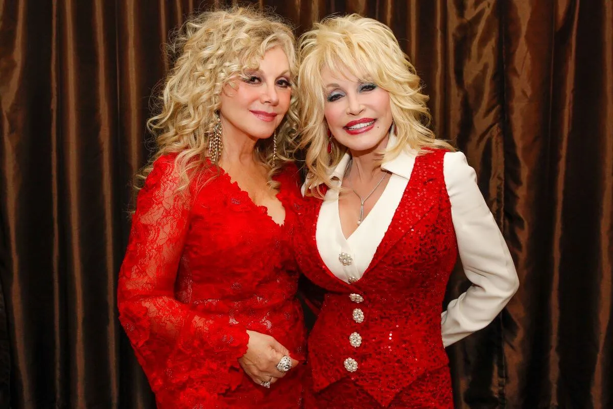 Stella Parton wears a red shirt with bell sleeves and Dolly Parton wears a red vest and white collared shirts. They stand with their arms around each other.