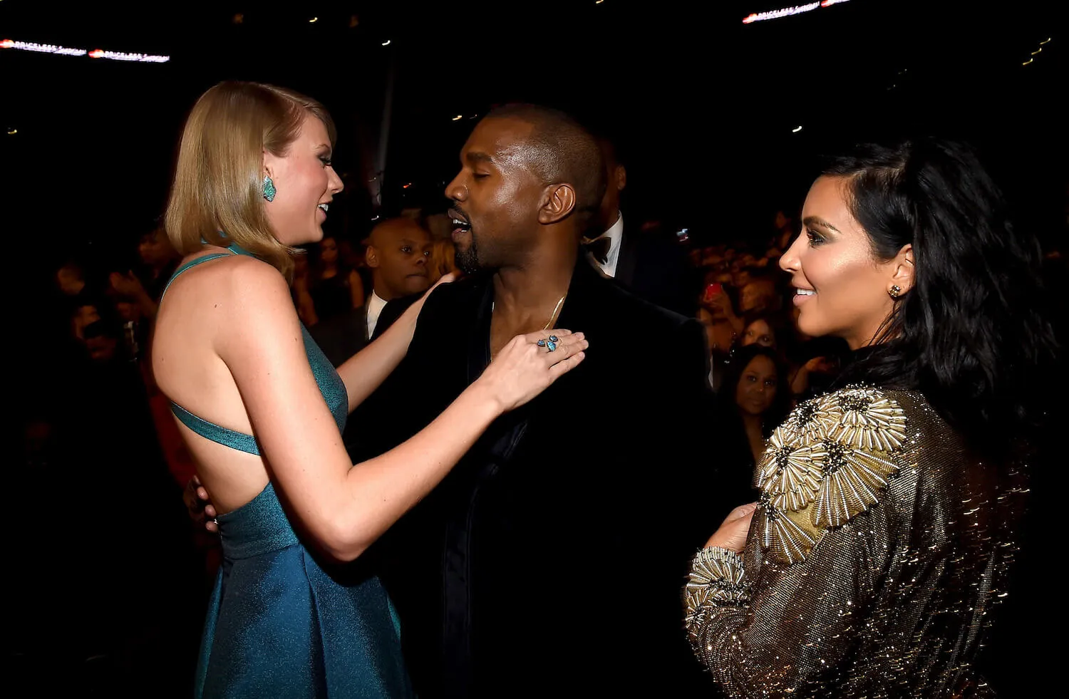 Taylor Swift with her hands on Kanye West's shoulders while talking to him at the 2015 Grammy Awards. Kim Kardashian is next to them. 