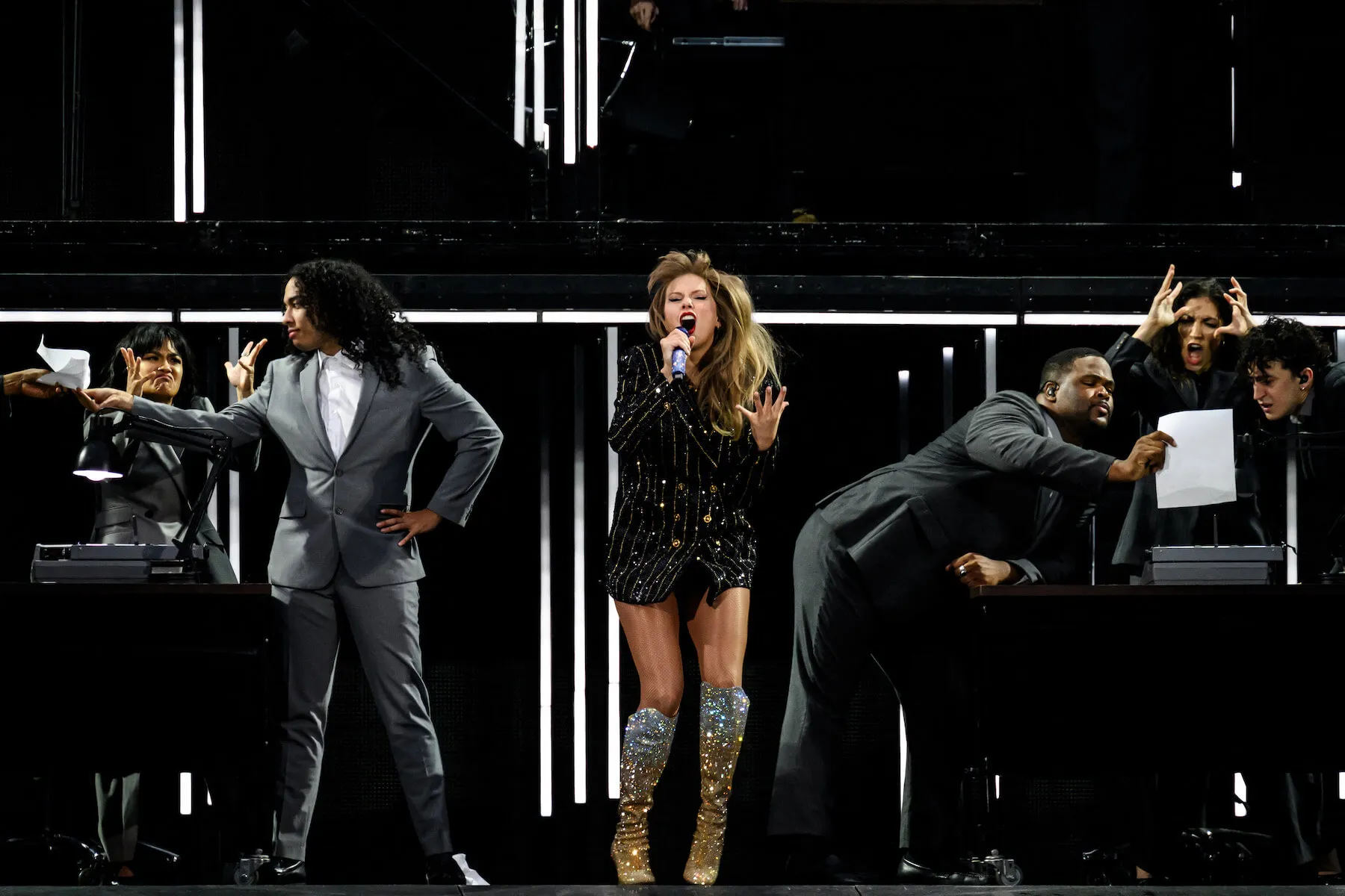 Taylor Swift wearing black and singing on stage in 'The Eras Tour' with male backup dancers in suits on either side of her