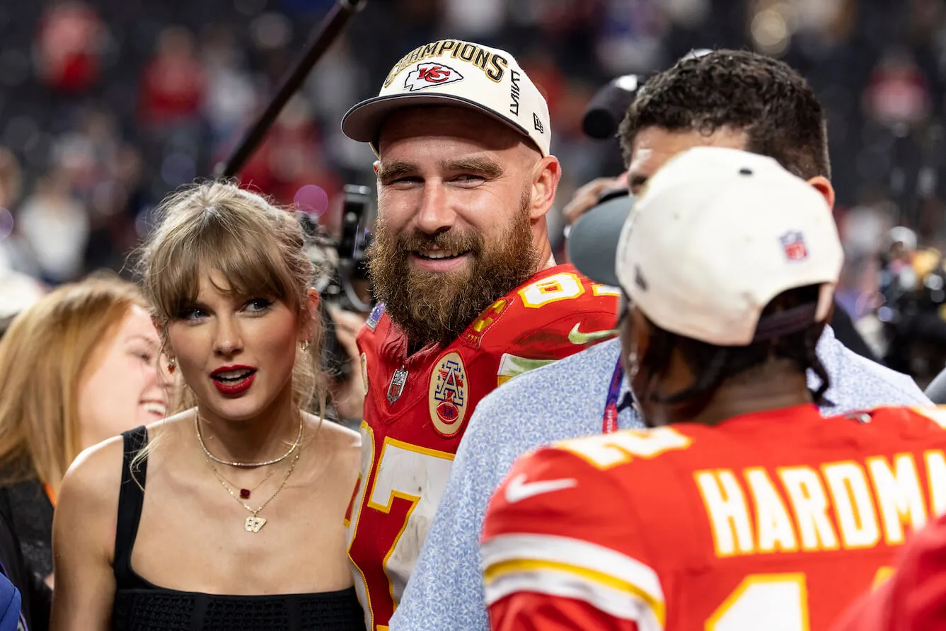 Taylor Swift standing next to Travis Kelce on the field in a crowd after a Kansas City Chiefs game