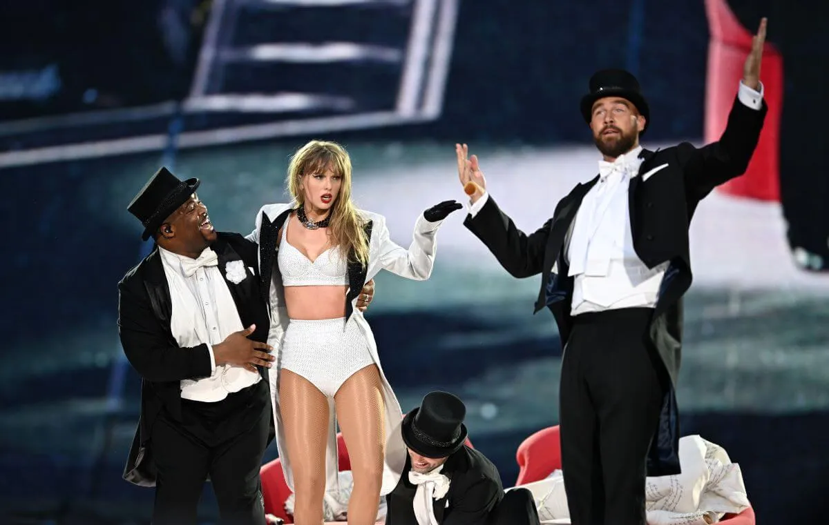 Taylor Swift stands onstage with backup dancers and Travis Kelce. Kelce and the dancers wear top hats and suits.