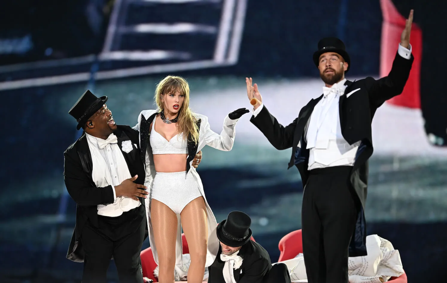 Taylor Swift in a white two-piece outfit on stage during 'The Eras Tour' in London with Travis Kelce in a suit and top hat next to her.