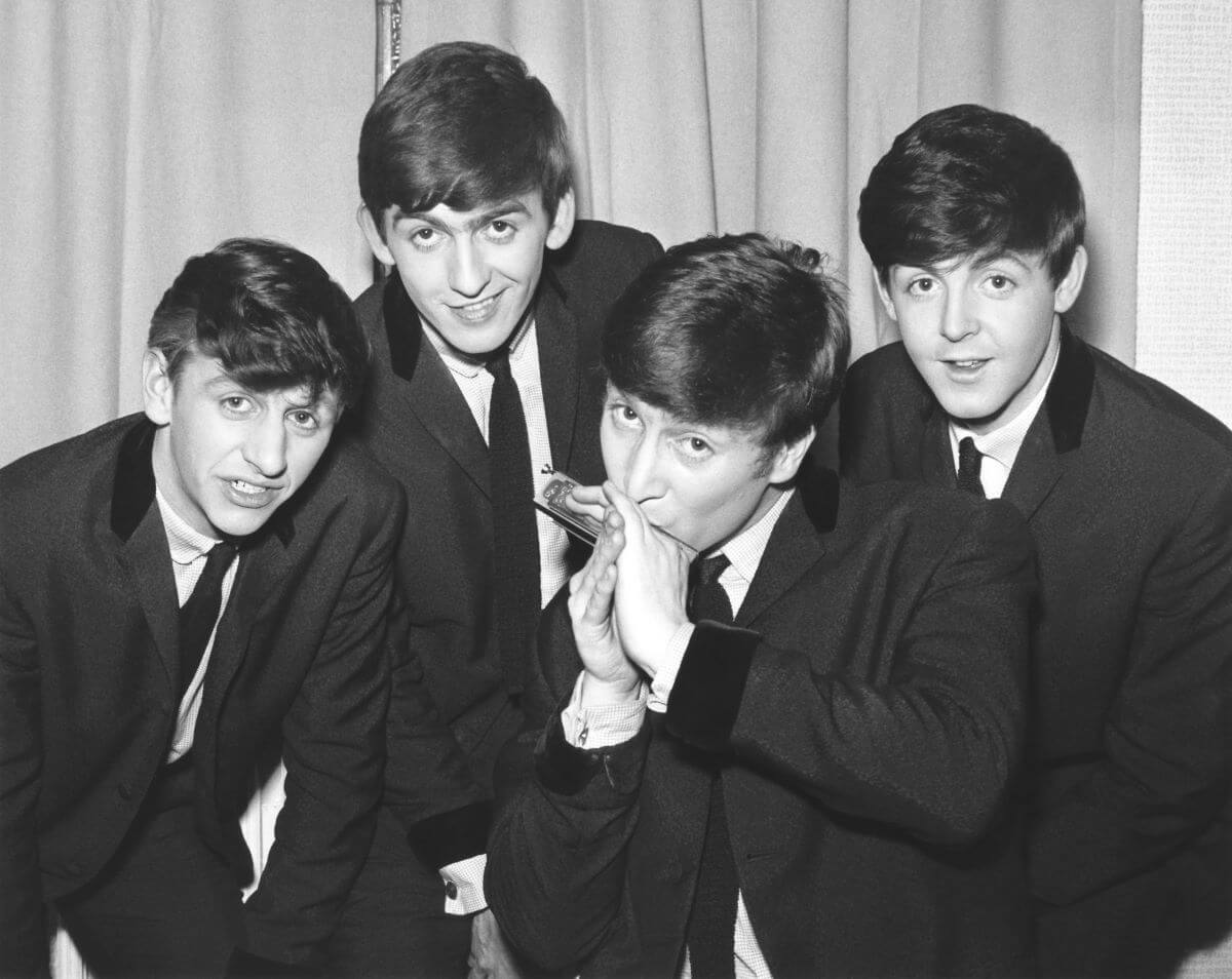 A black and white picture of The Beatles wearing suits and leaning close together. John Lennon plays the harmonica.
