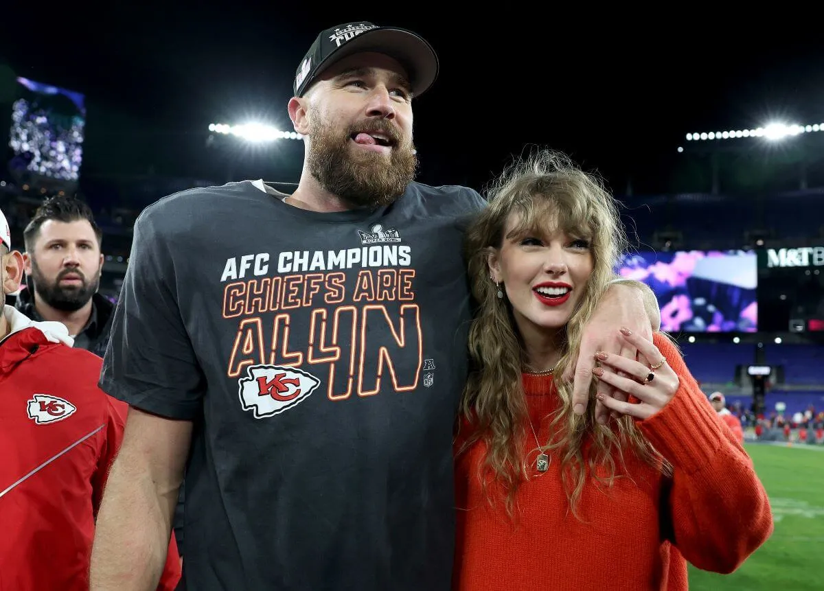 Travis Kelce wears a t-shirt, baseball hat, and football pads. He stands with his arm around Taylor Swift, who wears a red sweater.