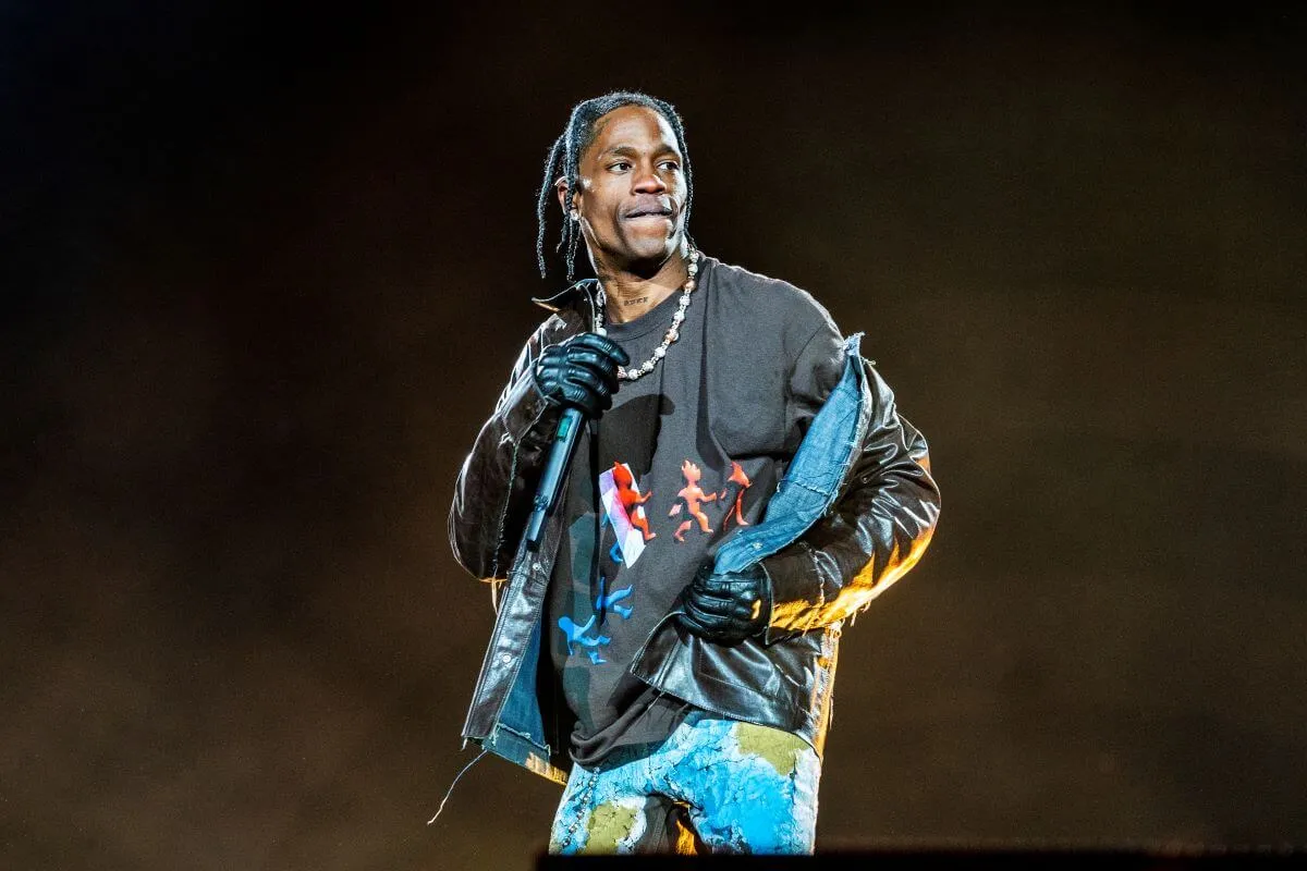 Travis Scott wears a leather jacket and gloves. He holds a microphone. 