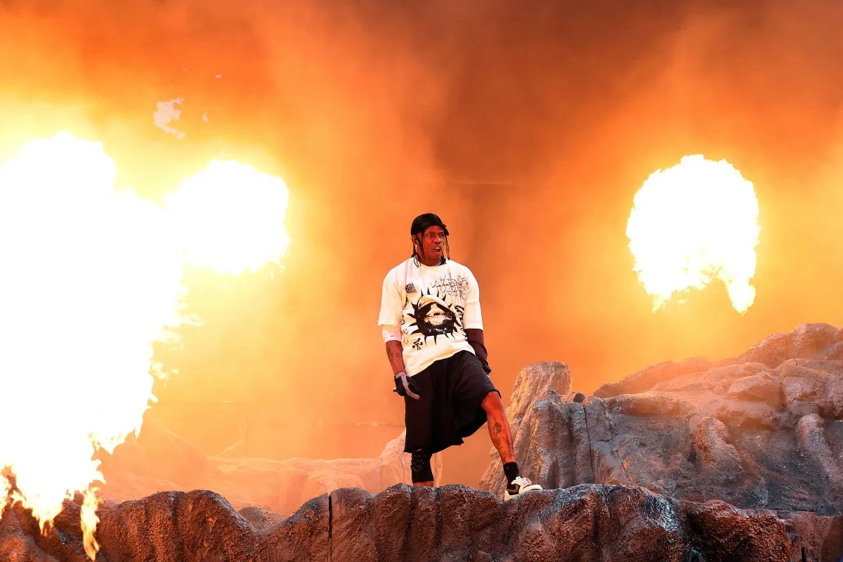 Travis Scott stands on a stage while pyrotechnics go off behind him.