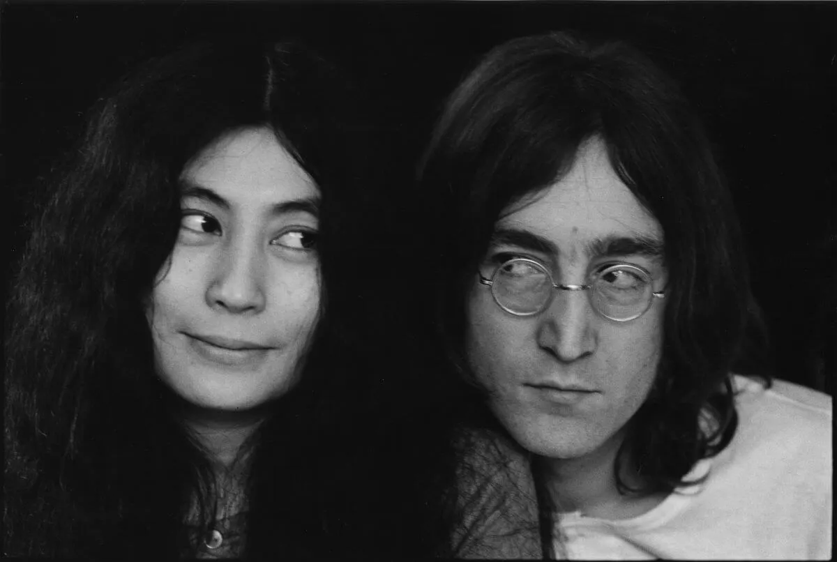 A black and white picture of Yoko Ono and John Lennon looking sideways at each other. He wears glasses.