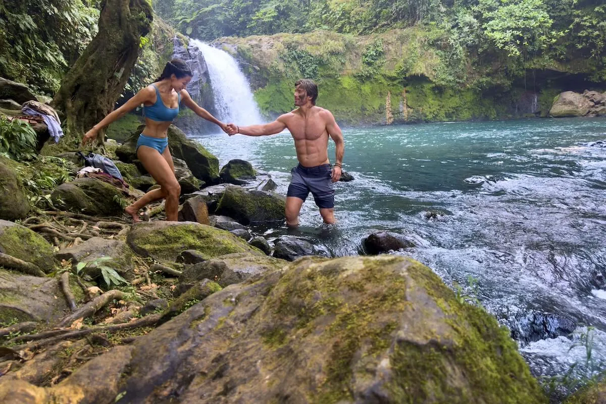 A couple scrambling on rocks in front of a waterfall in the Hallmark movie 'A Costa Rican Wedding'