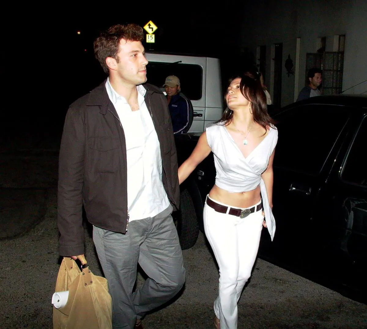 Ben Affleck and Jennifer Lopez hold hands while walking outside. She wears a white shirt and pants and he wears a white shirt and black jacket.