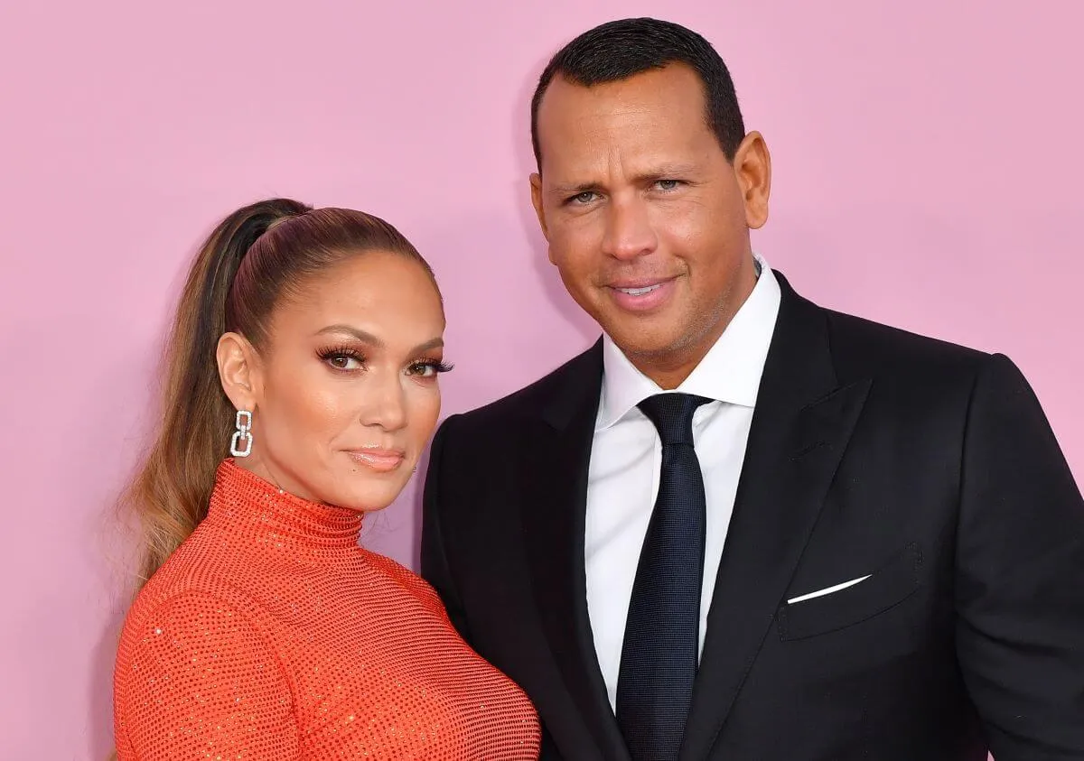 Jennifer Lopez wears orange and stands with Alex Rodriguez, who wears a suit. They stand in front of a pink wall.
