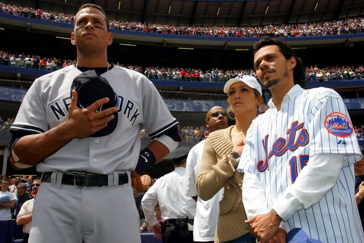 Alex Rodriguez, Jennifer Lopez, and Marc Anthony stand together at a Yankees game. Rodriguez wears his uniform and holds his hat to his chest.